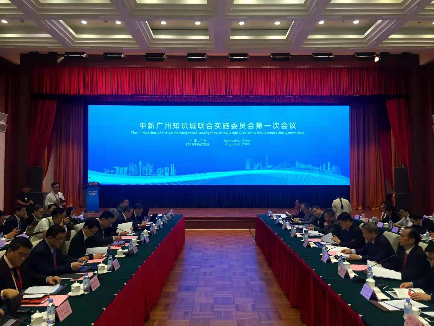 Meeting room with rows of people, stage with large screen, and red curtains.